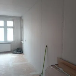 Pok&oacute;j w trakcie montażu płyt kartonowo-gipsowych na ścianach i suficie, widoczne łączenia płyt, na podłodze płyty OSB, przy ścianie oparta zielona rurka, obok stos płyt