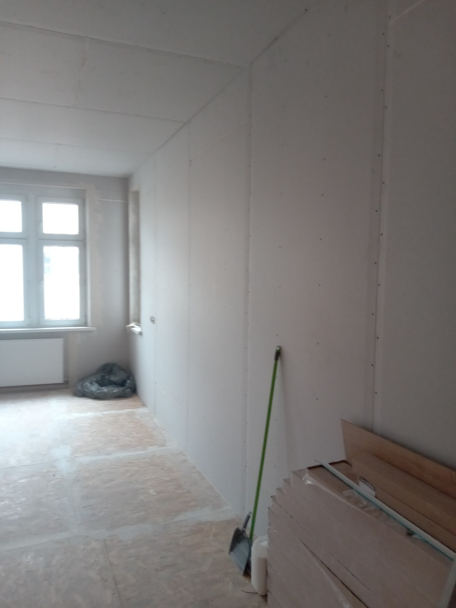 Pokój w trakcie montażu płyt kartonowo-gipsowych na ścianach i suficie, widoczne łączenia płyt, na podłodze płyty OSB, przy ścianie oparta zielona rurka, obok stos płyt