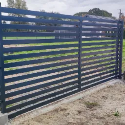 Nowoczesna, pozioma brama metalowa w kolorze grafitowym z przylegającym ogrodzeniem panelowym, widoczna na tle domu jednorodzinnego i trawnika.