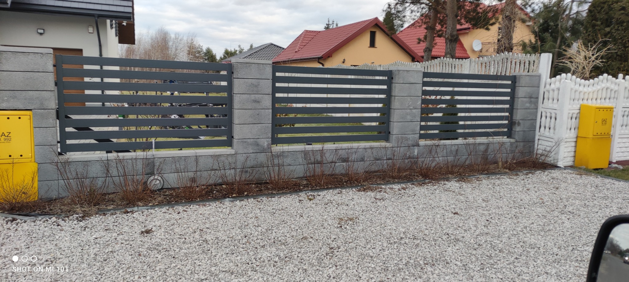 Nowoczesne ogrodzenie z szarego metalu z poziomymi przęsłami i betonowymi słupkami, widok z zewnątrz na posesję z białym płotem i żółtą skrzynką gazową.