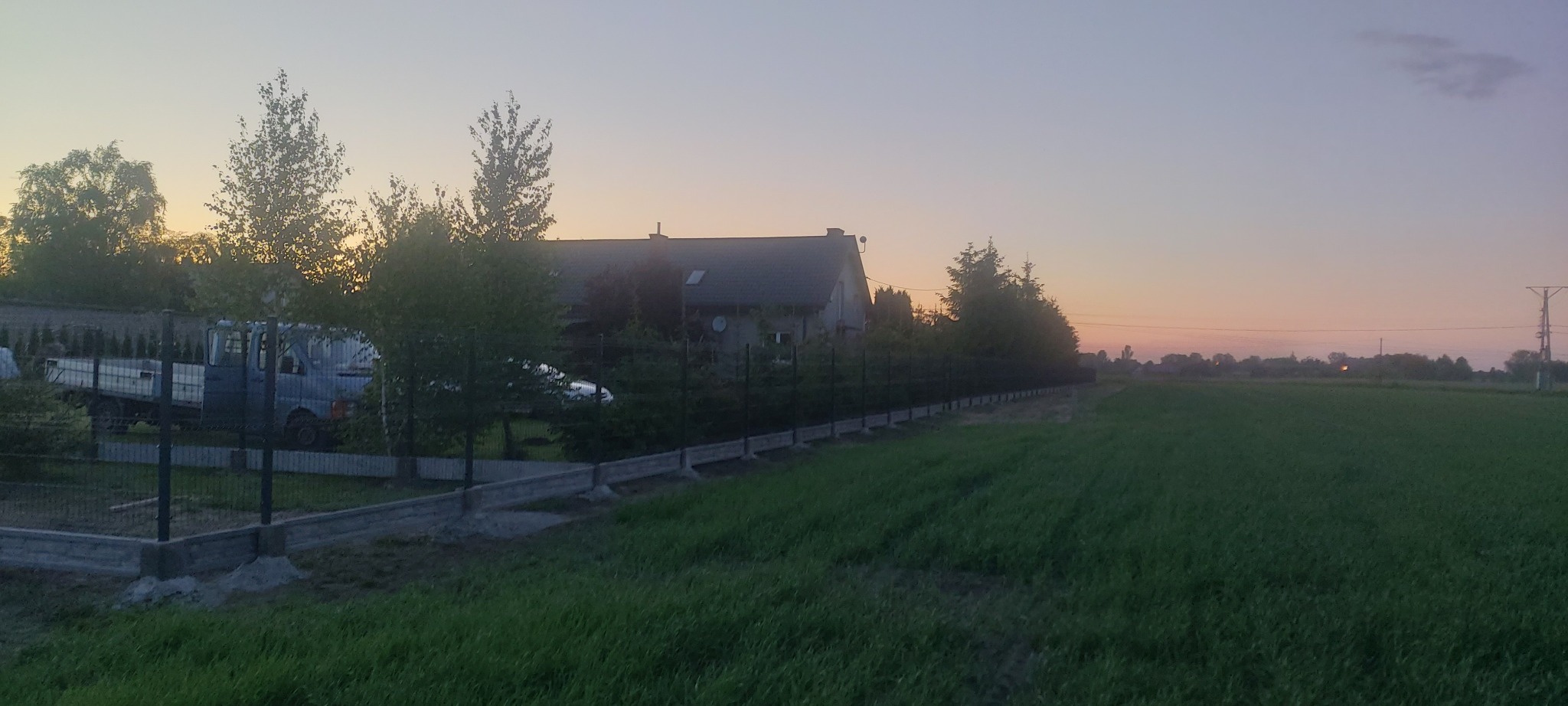 Nowo postawione ogrodzenie z paneli na betonowej podmurówce, widoczne na tle domu i pola o zmierzchu.