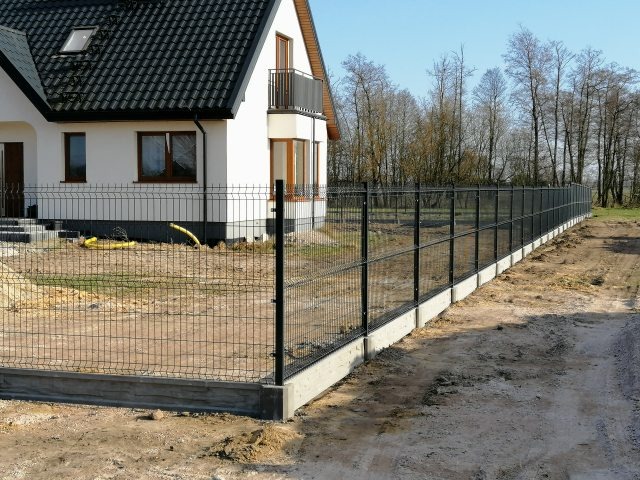 Nowoczesne ogrodzenie z paneli zgrzewanych na betonowej podmurówce, otaczające posesję z białym domem z balkonem, widok z zewnątrz.