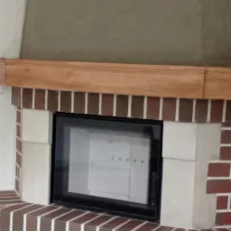 Nowoczesny kominek obudowany cegłą klinkierową z elementami z jasnego kamienia, z drewnianą półką nad wkładem kominkowym i otworem wentylacyjnym w górnej części obudowy.