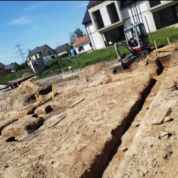 ARTEAM Grzegorz Argier - Mini koparka wykonuje wykop pod fundamenty domu jednorodzinnego na piaszczystym terenie, widoczne rury gazowe i zbrojenie.