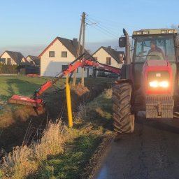 ARTEAM Grzegorz Argier - Czerwony traktor z wysięgnikiem i kosiarką rotacyjną przy rowie melioracyjnym, w tle domy jednorodzinne i błękitne niebo.