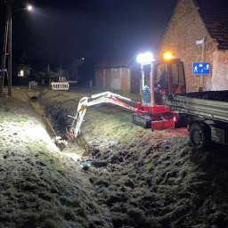 ARTEAM Grzegorz Argier - Mini koparka z operatorem oświetlona reflektorami wykopuje wąski rów w ziemi w nocy, obok stoi ciężarówka, w tle budynek z cegły i znak drogowy.