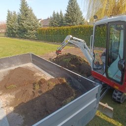 ARTEAM Grzegorz Argier - Mini koparka Takeuchi TB216 nabiera ziemię do metalowej przyczepy na tle zielonego trawnika i żywopłotu.