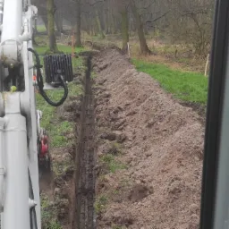 Wąski wykop w ziemi, wykonany prawdopodobnie koparką, widok z kabiny operatora na tle drzew i zielonej trawy.