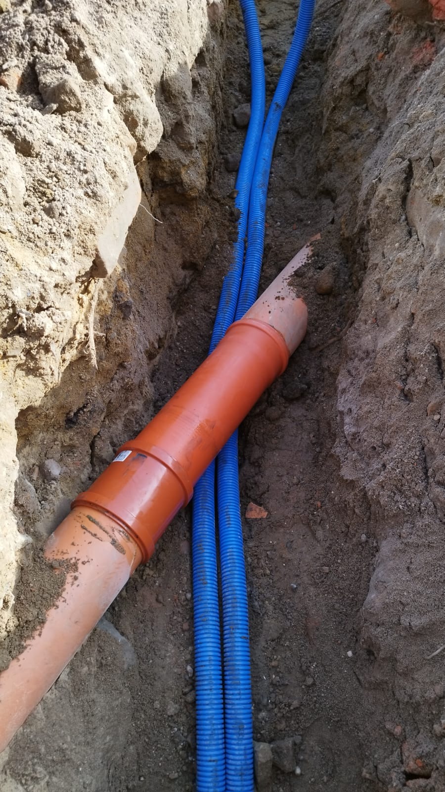 Wykop w ziemi z pomarańczową rurą kanalizacyjną i dwiema niebieskimi rurami karbowanymi ułożonymi wzdłuż wykopu.