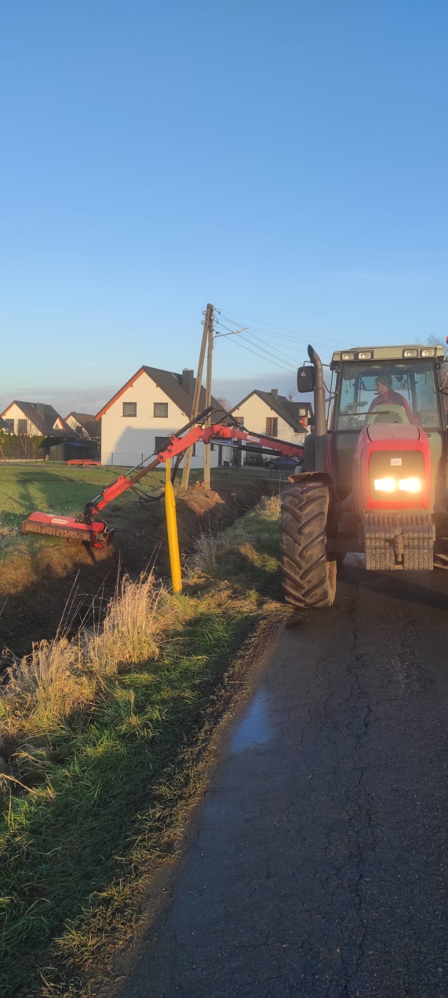 Czerwony traktor z wysięgnikiem i kosiarką rotacyjną przy rowie melioracyjnym, w tle domy jednorodzinne i błękitne niebo.
