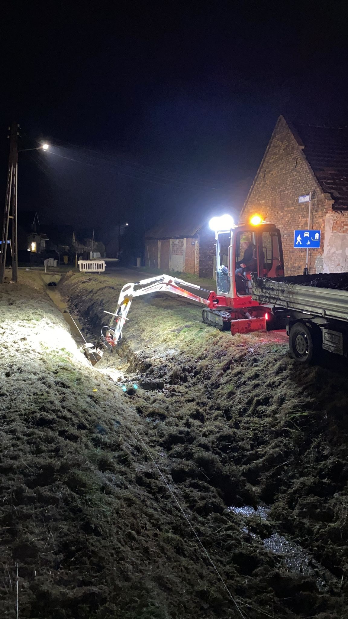 Mini koparka z operatorem oświetlona reflektorami wykopuje wąski rów w ziemi w nocy, obok stoi ciężarówka, w tle budynek z cegły i znak drogowy.