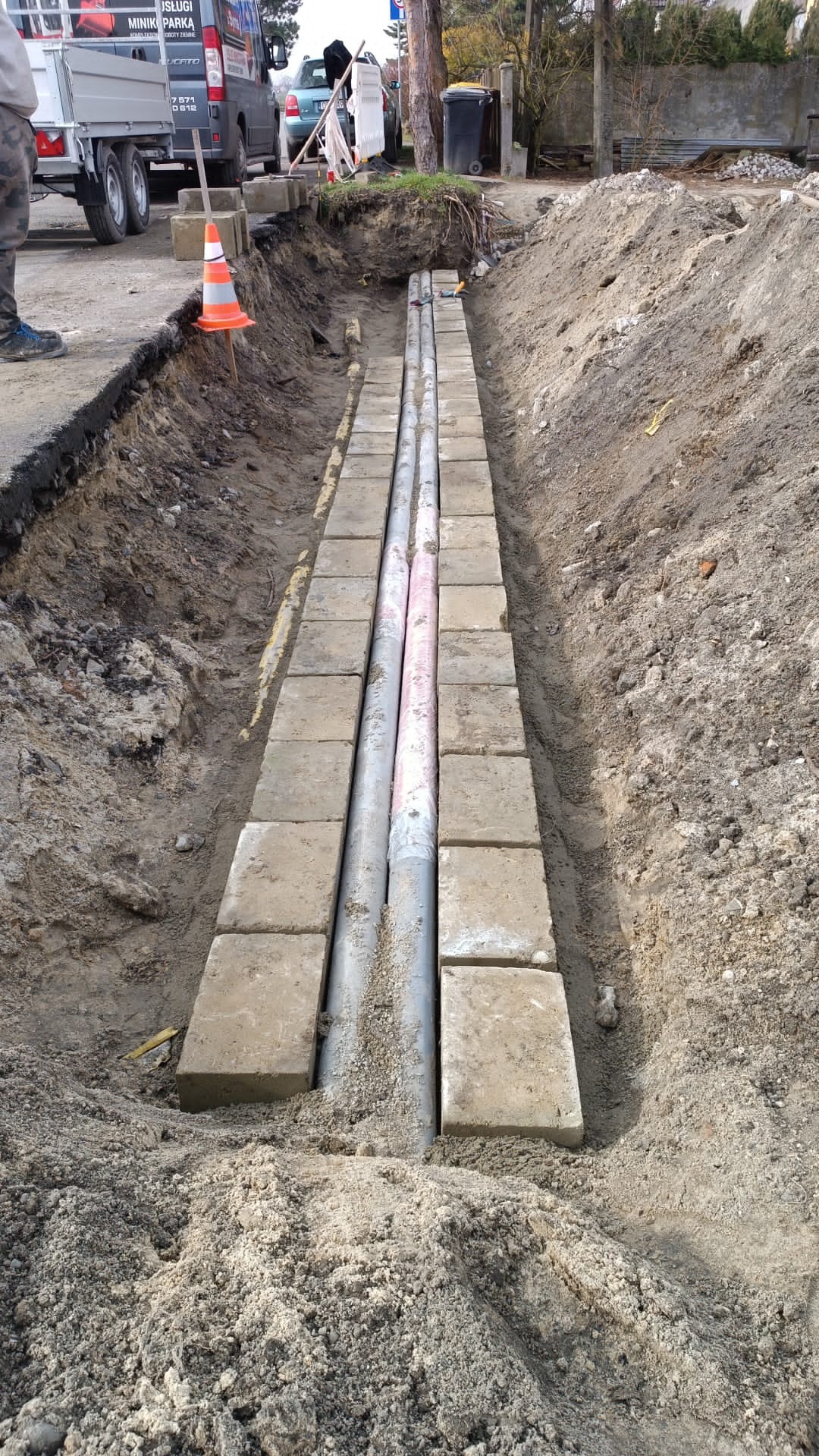 Wykop w ziemi z ułożonymi rurami i obłożony betonowymi płytami, widoczny fragment drogi i samochód dostawczy w tle.