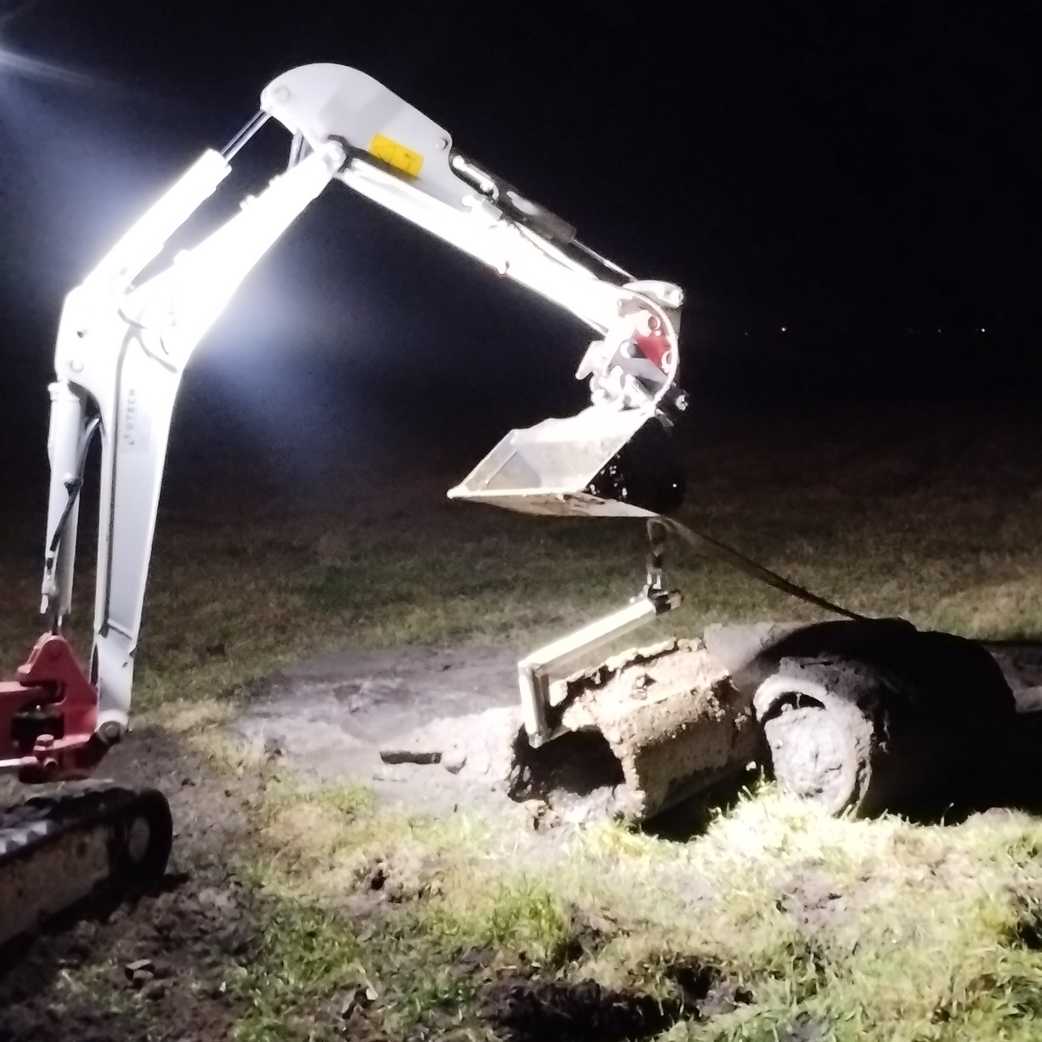 Mini koparka oświetlona reflektorem przy wykopanym fragmencie rury kanalizacyjnej w ciemnościach.