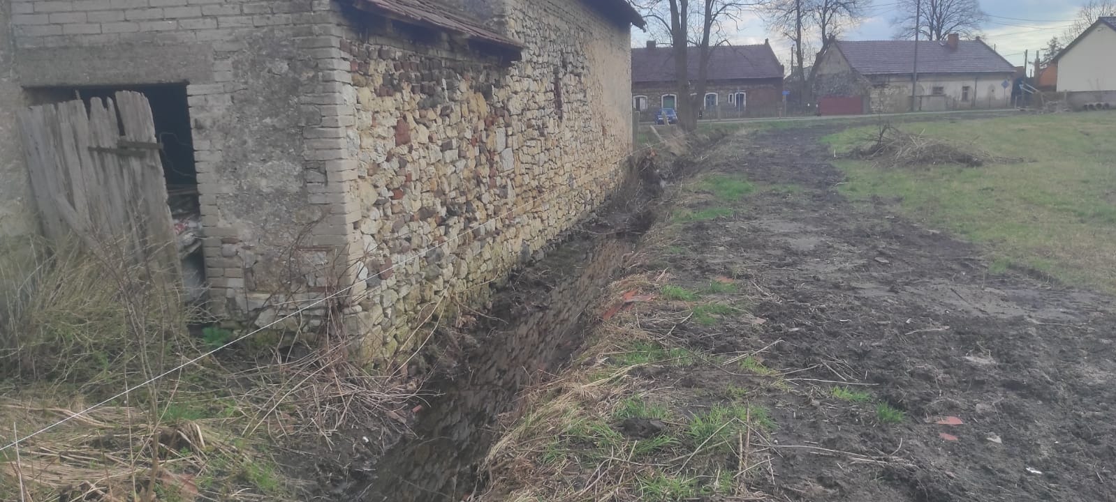 Wykonany drenaż wzdłuż starego, kamiennego budynku gospodarczego z otwartymi wrotami, widoczny wykop w ziemi.