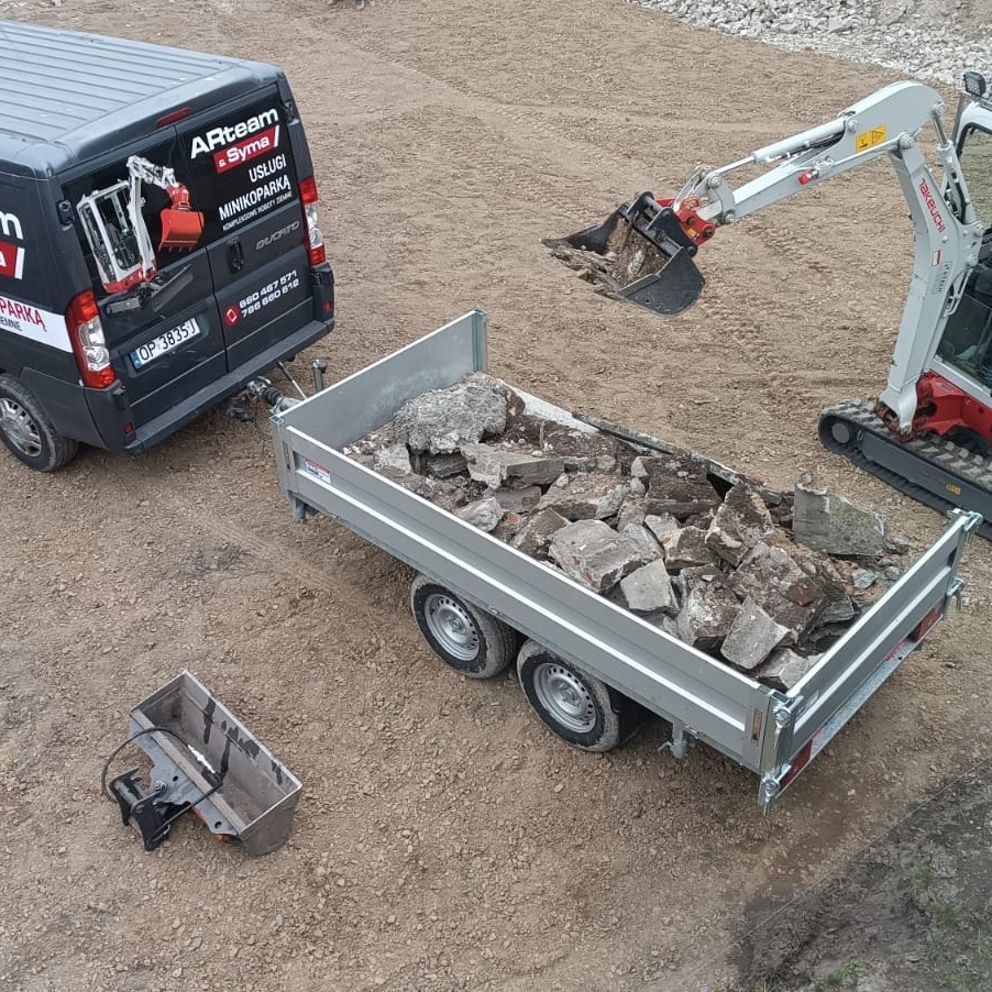 Minikoparka Takeuchi nabiera gruz do przyczepy, którą ciągnie czarny bus z logo firmy ARteam oferującej usługi minikoparką, w tle przygotowany teren pod budowę.