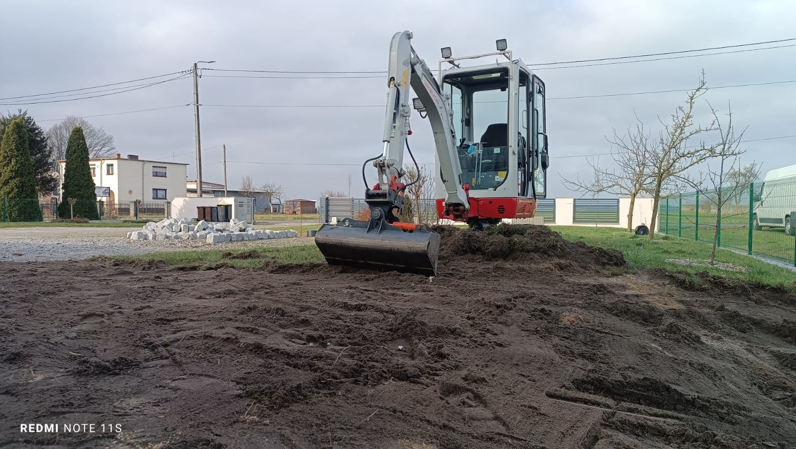Mała koparka Takeuchi z czarną łyżką na nierównym, mokrym terenie; w tle budynek mieszkalny, sterta kostki brukowej i pochmurne niebo.