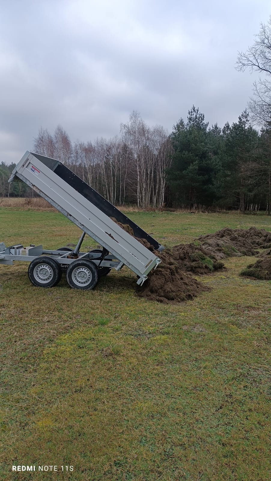Przyczepa wywrotka z podniesioną skrzynią ładunkową, wysypująca ziemię z darnią na łąkę, w tle las i pochmurne niebo.