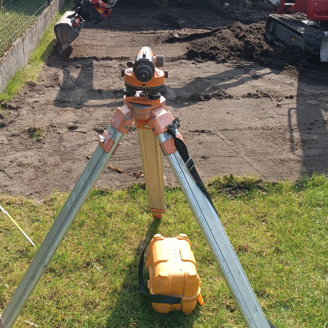 Niwelator optyczny na statywie, żółta skrzynka obok, w tle wykop ziemny i minikoparka w trakcie niwelacji terenu.