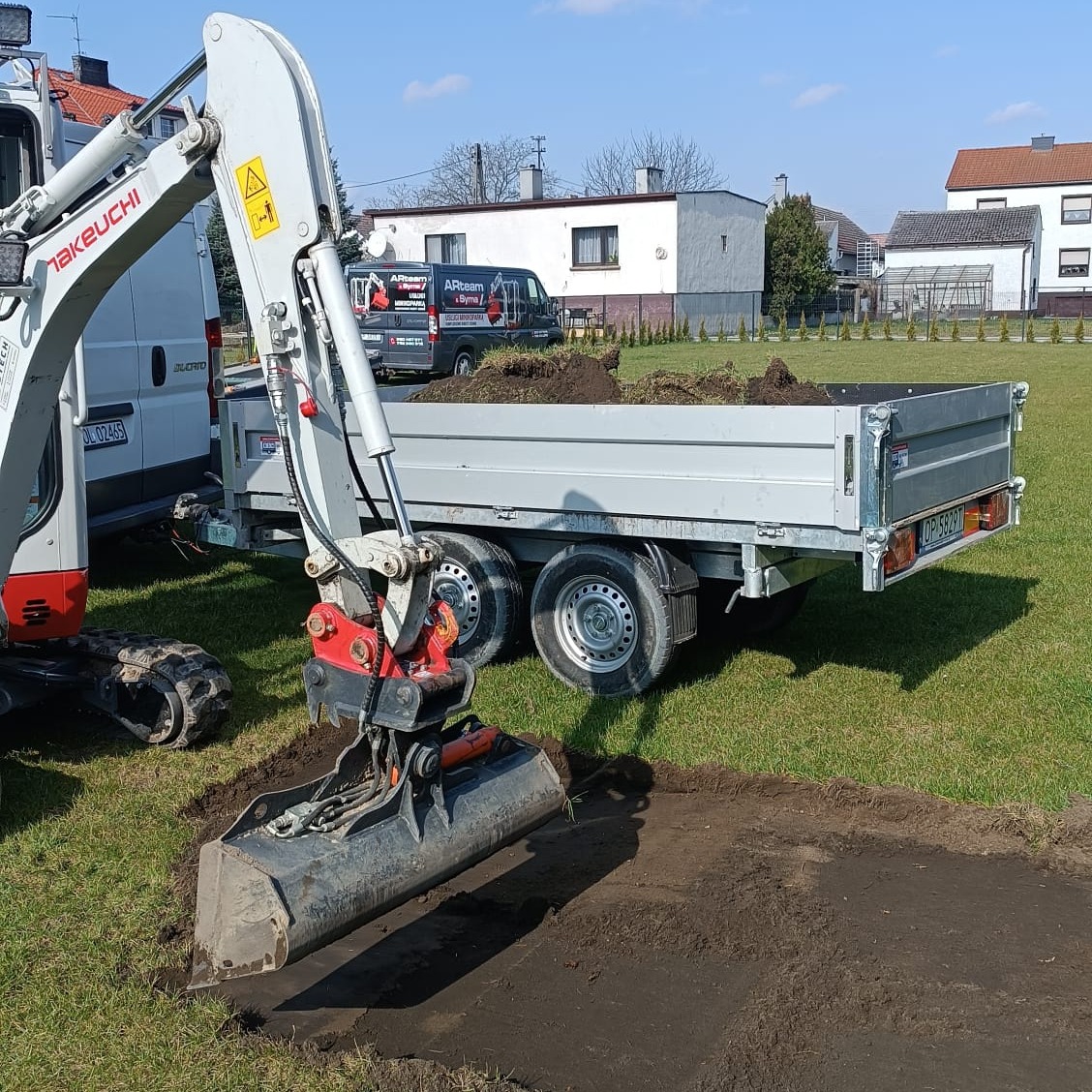 Mini koparka Takeuchi z łyżką w trakcie prac ziemnych na trawniku; przyczepa z ładunkiem ziemi w tle oraz zaparkowany bus firmy budowlanej.