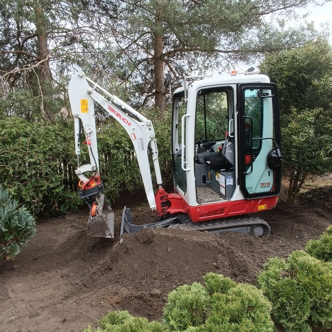 Mała, czerwono-biała koparka Takeuchi TB216 na gąsienicach, zagłębiająca łyżkę w ziemi na tle zielonych krzewów i drzew.