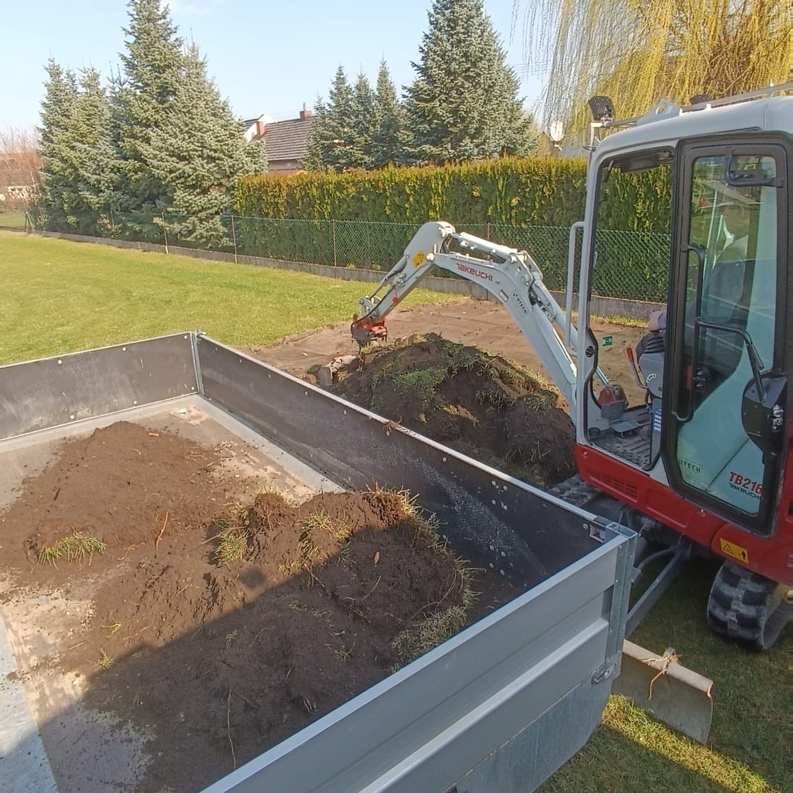 Mini koparka Takeuchi TB216 nabiera ziemię do metalowej przyczepy na tle zielonego trawnika i żywopłotu.