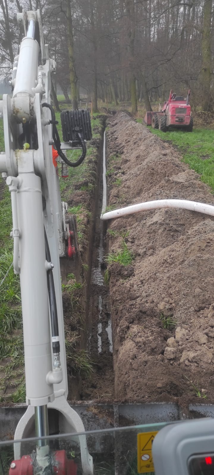 Wykopany wąski rów w ziemi z ułożoną białą rurą, widziany z perspektywy operatora koparki, z tyłu widoczny czerwony mini-ładowarka na tle drzew.