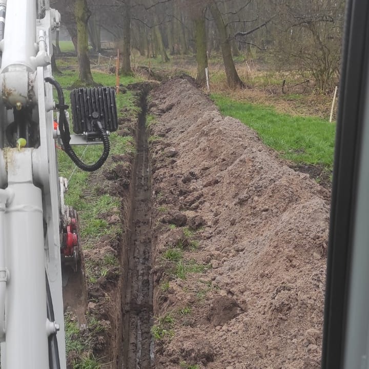 Wąski wykop w ziemi, wykonany prawdopodobnie koparką, widok z kabiny operatora na tle drzew i zielonej trawy.