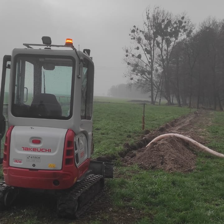 Mała koparka Takeuchi na polu, świeżo wykopany rów z widoczną rurą, pochmurny dzień.