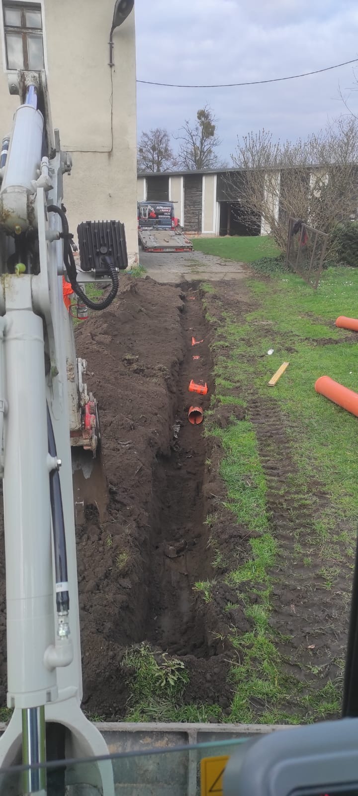 Wykopany rów pod rury kanalizacyjne na trawniku, widok z kabiny koparki w pochmurny dzień.