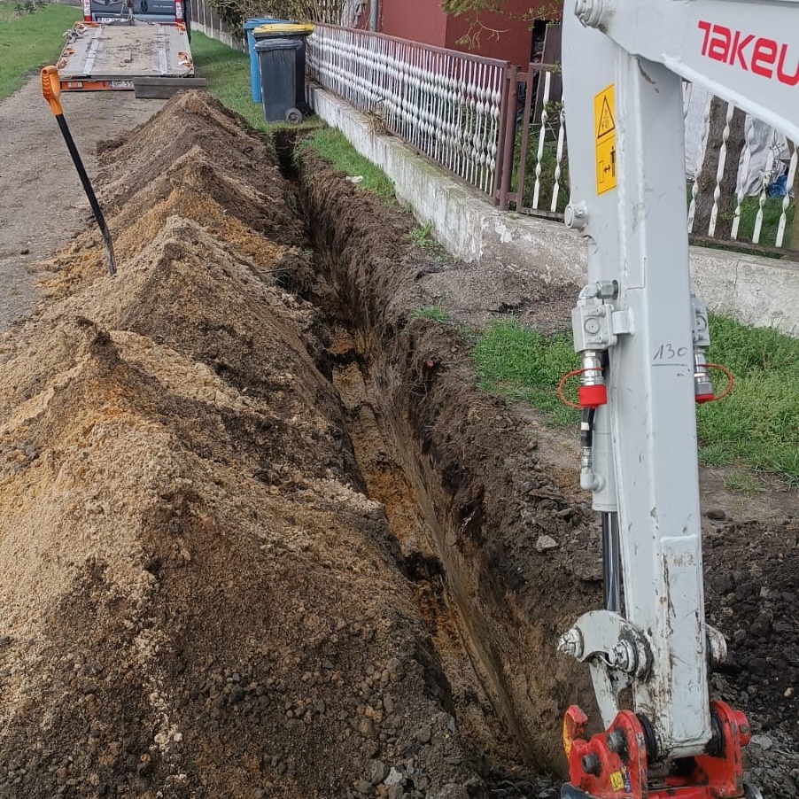 Wykopany wąski rów w ziemi, obok sterta piasku i łopata, w tle widoczny fragment koparki Takeuchi i ogrodzenie posesji.