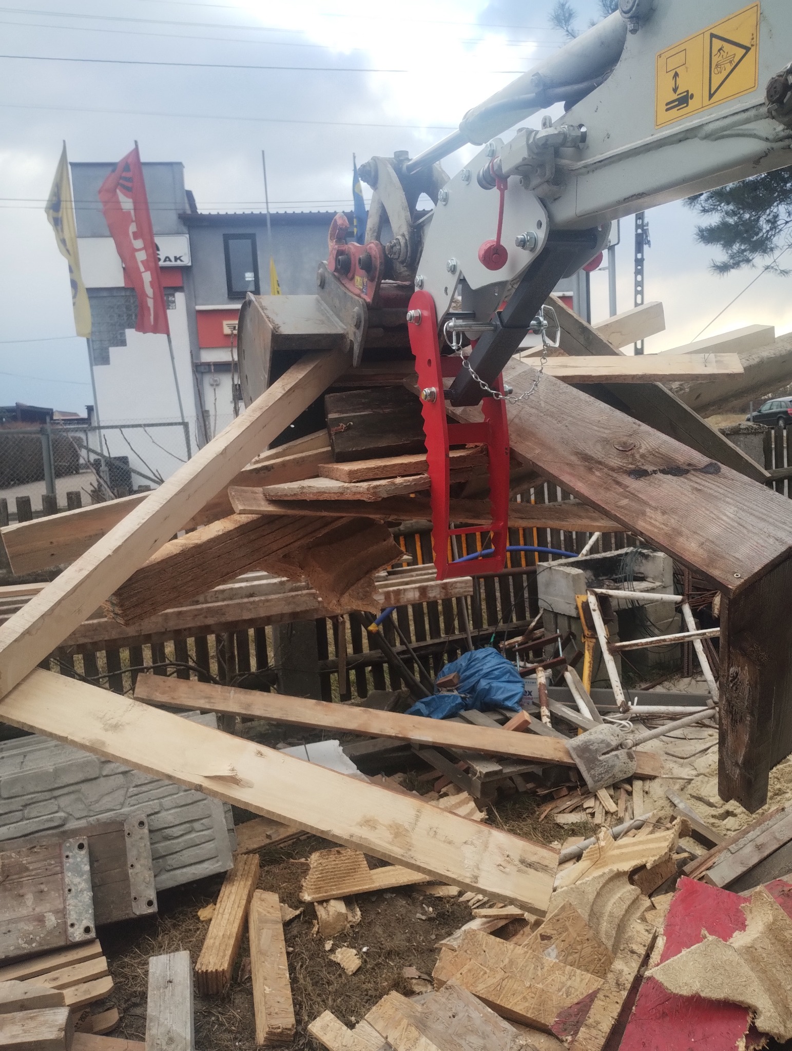 Hydrauliczny chwytak zamontowany na ramieniu koparki podnosi pozostałości drewnianej konstrukcji po rozbiórce, w tle budynek z reklamą i flagi.