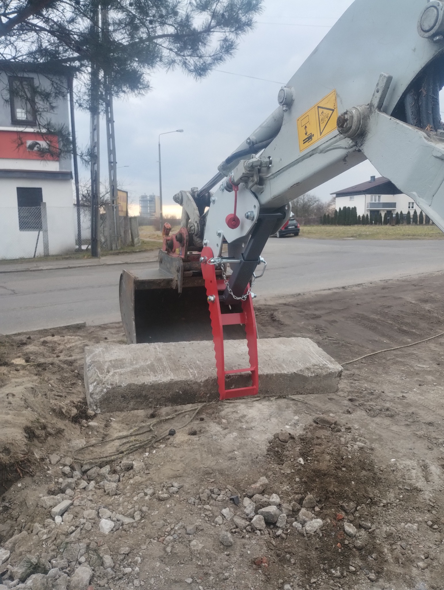 Koparka z czerwonym osprzętem podnosi betonowy blok na placu budowy, w tle budynek mieszkalny i ulica.