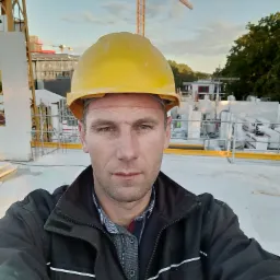 Autoportret budowlańca w żółtym kasku na tle budowy z białych bloczków i żurawia, uchwycony z niskiej perspektywy.