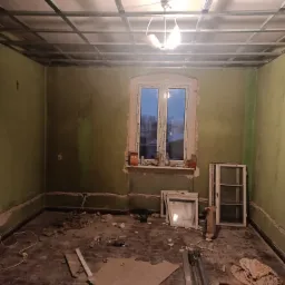 Wnętrze pokoju w trakcie remontu: zielone ściany, metalowa konstrukcja sufitu podwieszanego, stare okna, gruz na podłodze, widoczne kable elektryczne.