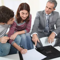 Młoda para podpisuje dokument na białym stole, doradca w szarym garniturze wskazuje palcem na formularz.