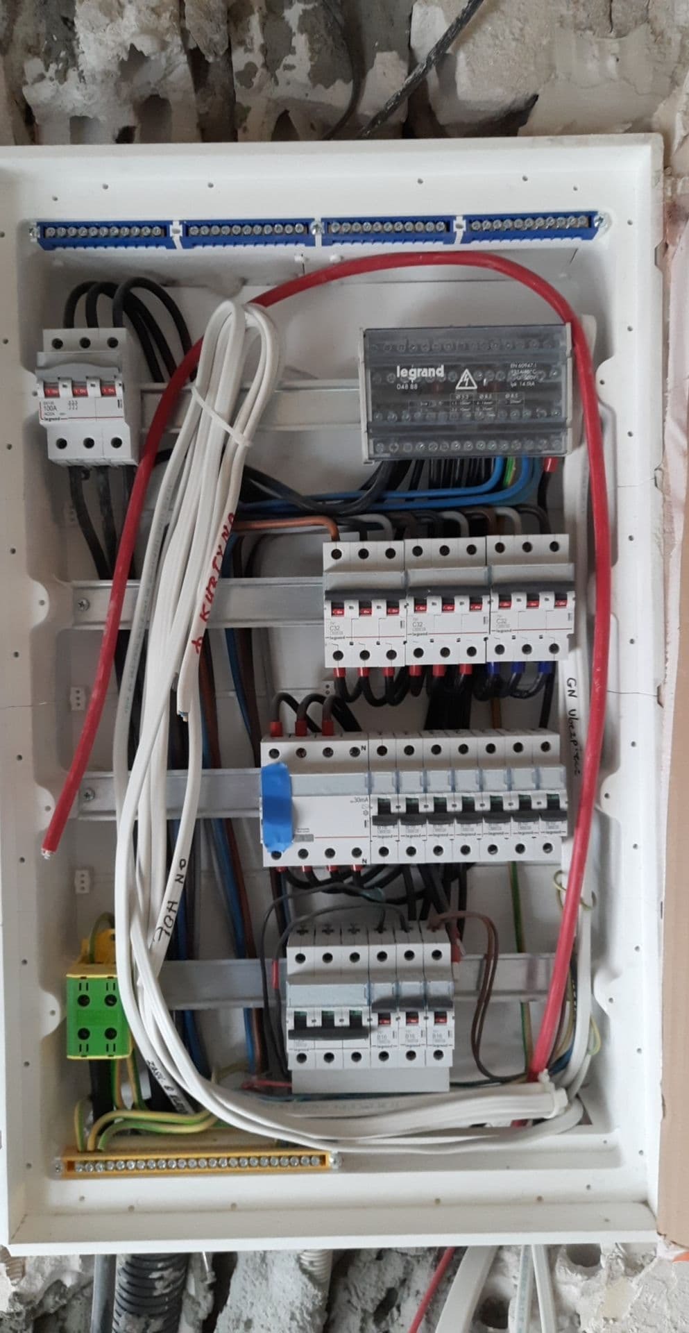 Otwarta skrzynka rozdzielcza z widocznymi bezpiecznikami, przewodami w kolorach białym, czerwonym i czarnym, zamontowana na surowej ścianie w trakcie remontu.