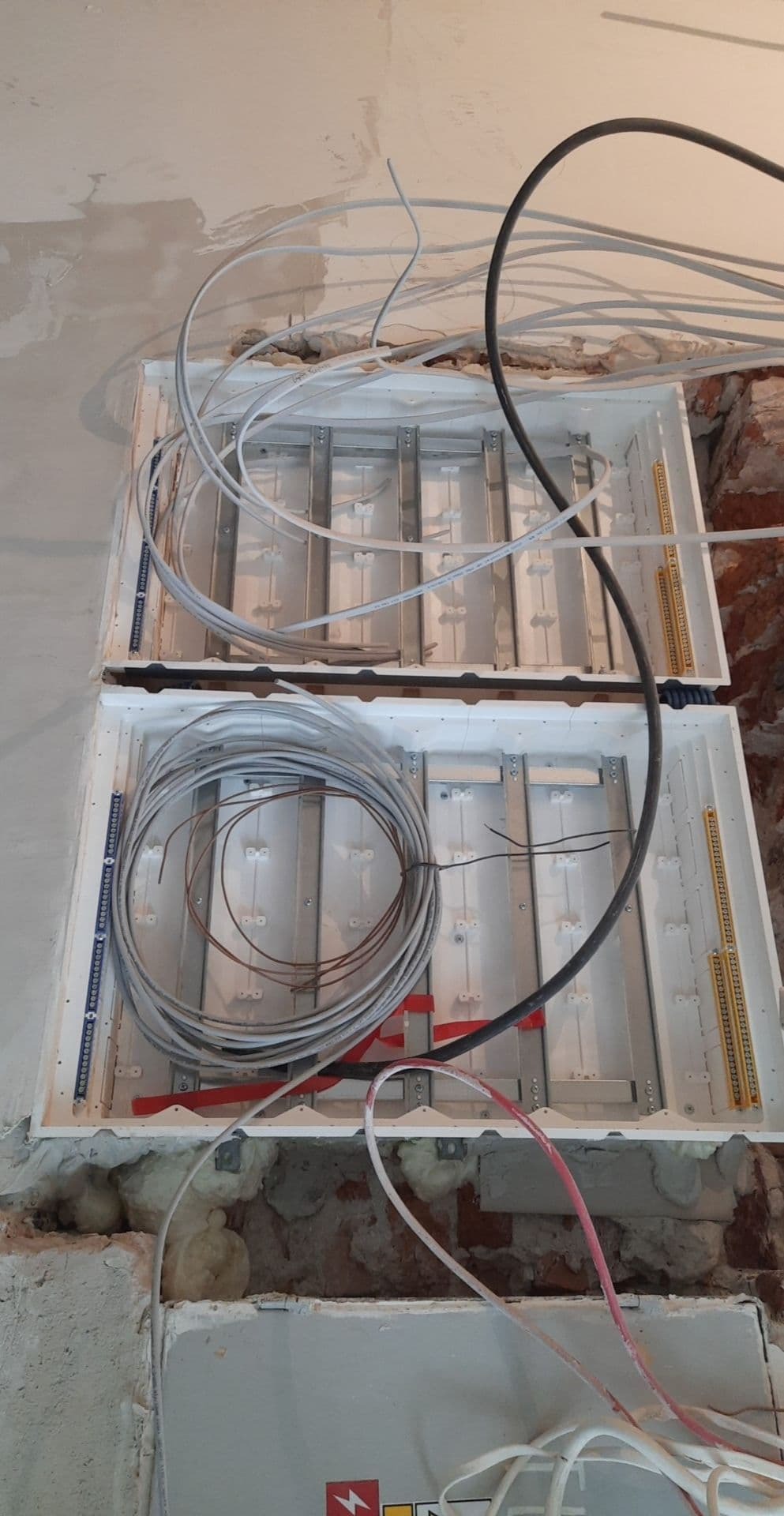 Dwa puste, białe panele elektryczne zamontowane w ścianie z widocznymi przewodami w różnych kolorach i grubościach, niektóre zwinięte w kłębek; ściana w stanie surowym z widocznymi cegłami i pianką...