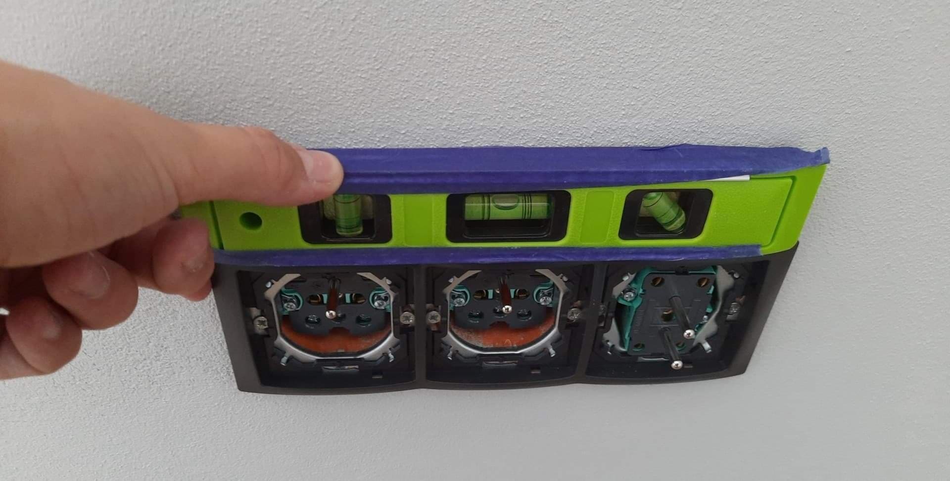 Poziomica przykładana do potrójnego gniazdka elektrycznego w trakcie montażu, widoczne przewody i elementy mocujące.