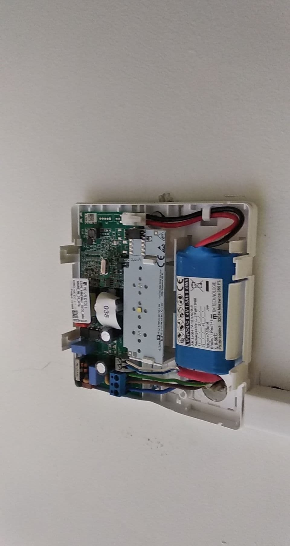 Otwarta obudowa urządzenia elektrycznego na ścianie, widoczne kable, płytka drukowana, bateria i złącza, z bliska.