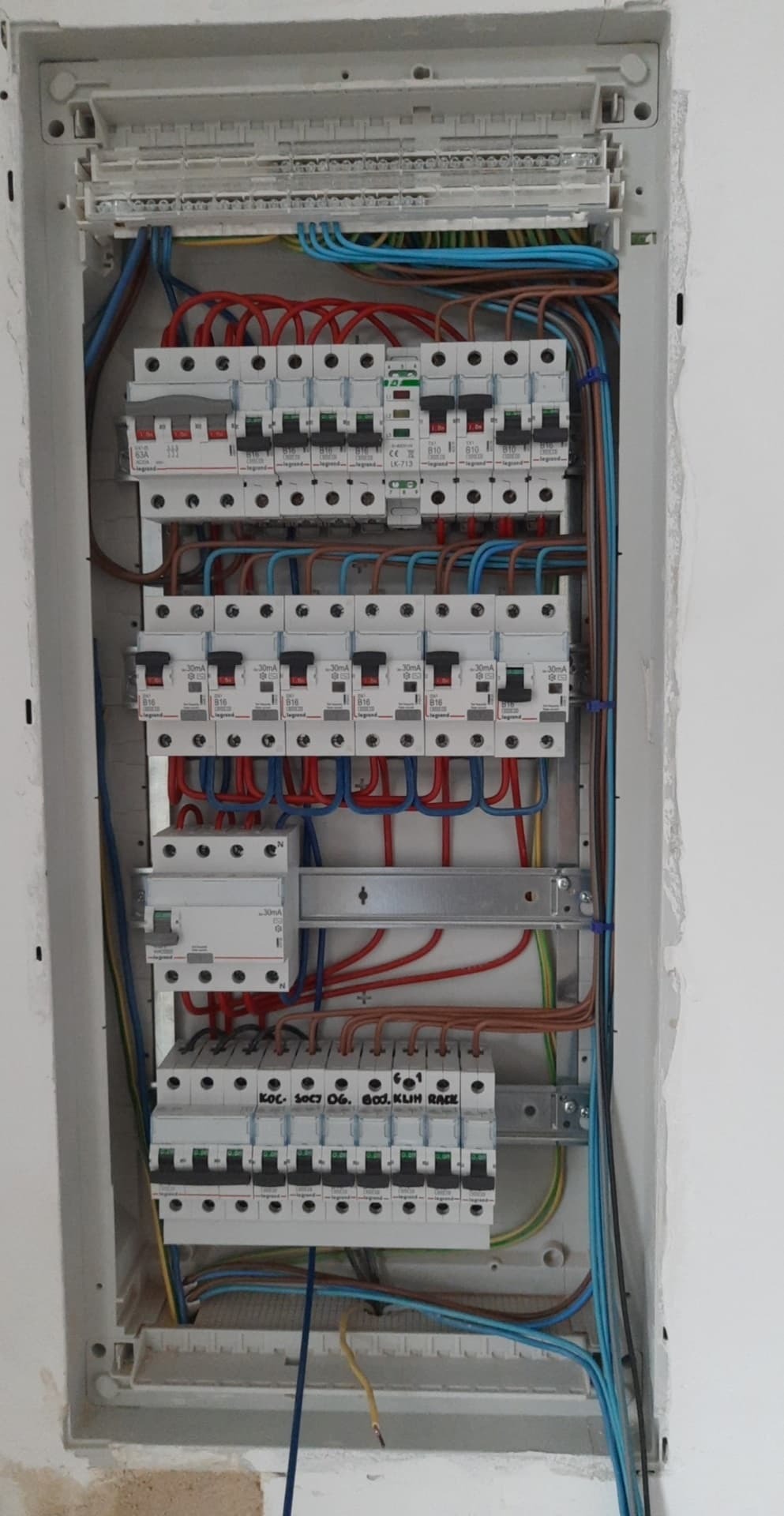 Otwarta skrzynka elektryczna z widocznymi bezpiecznikami, przewodami w kolorach niebieskim, czerwonym i brązowym, zamontowana na białej ścianie w trakcie instalacji.