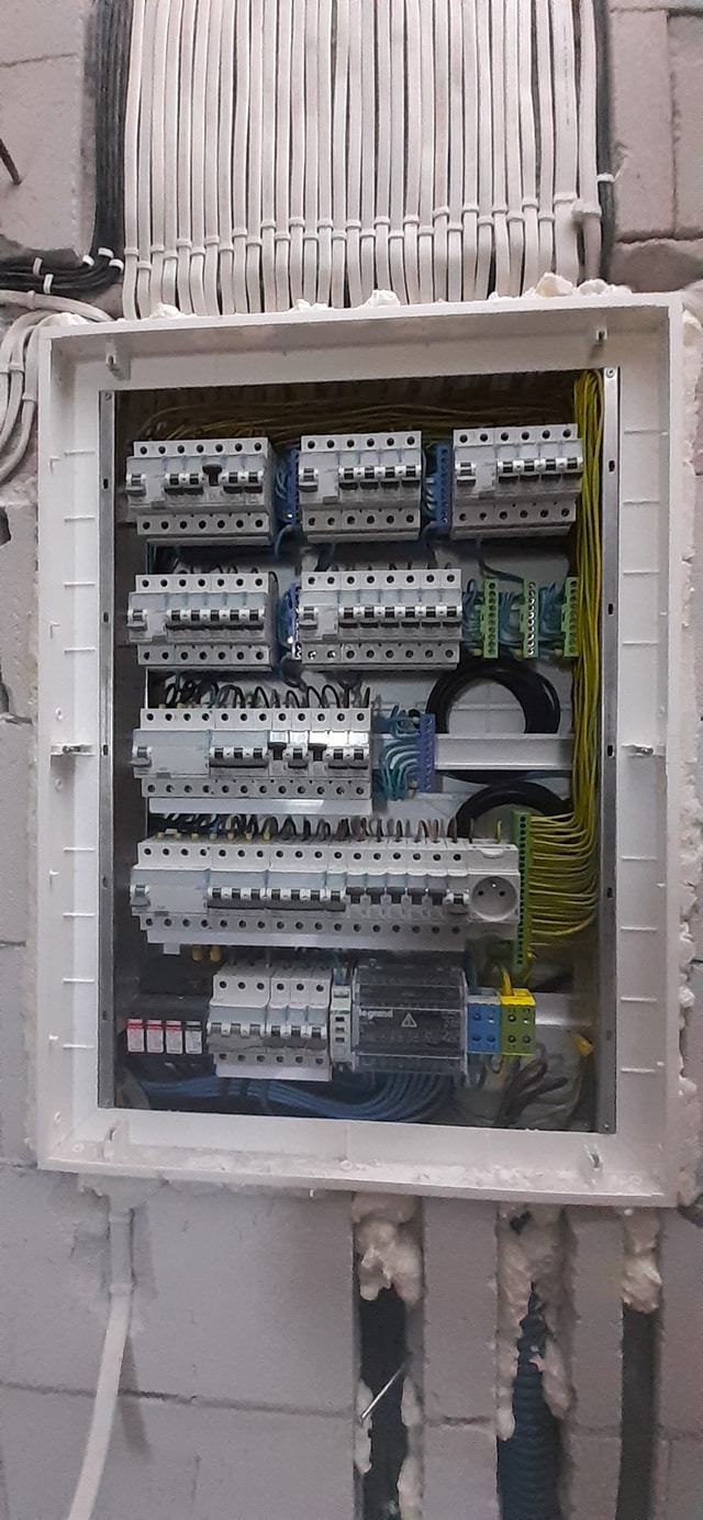 Rozdzielnica elektryczna z zabezpieczeniami, okablowaniem i gniazdem, zamontowana na ścianie z pustaków z widoczną pianką montażową.