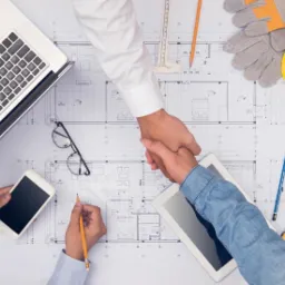 Uścisk dłoni nad planami architektonicznymi, laptopem, tabletem, smartfonem, okularami, żółtym kaskiem i narzędziami mierniczymi.