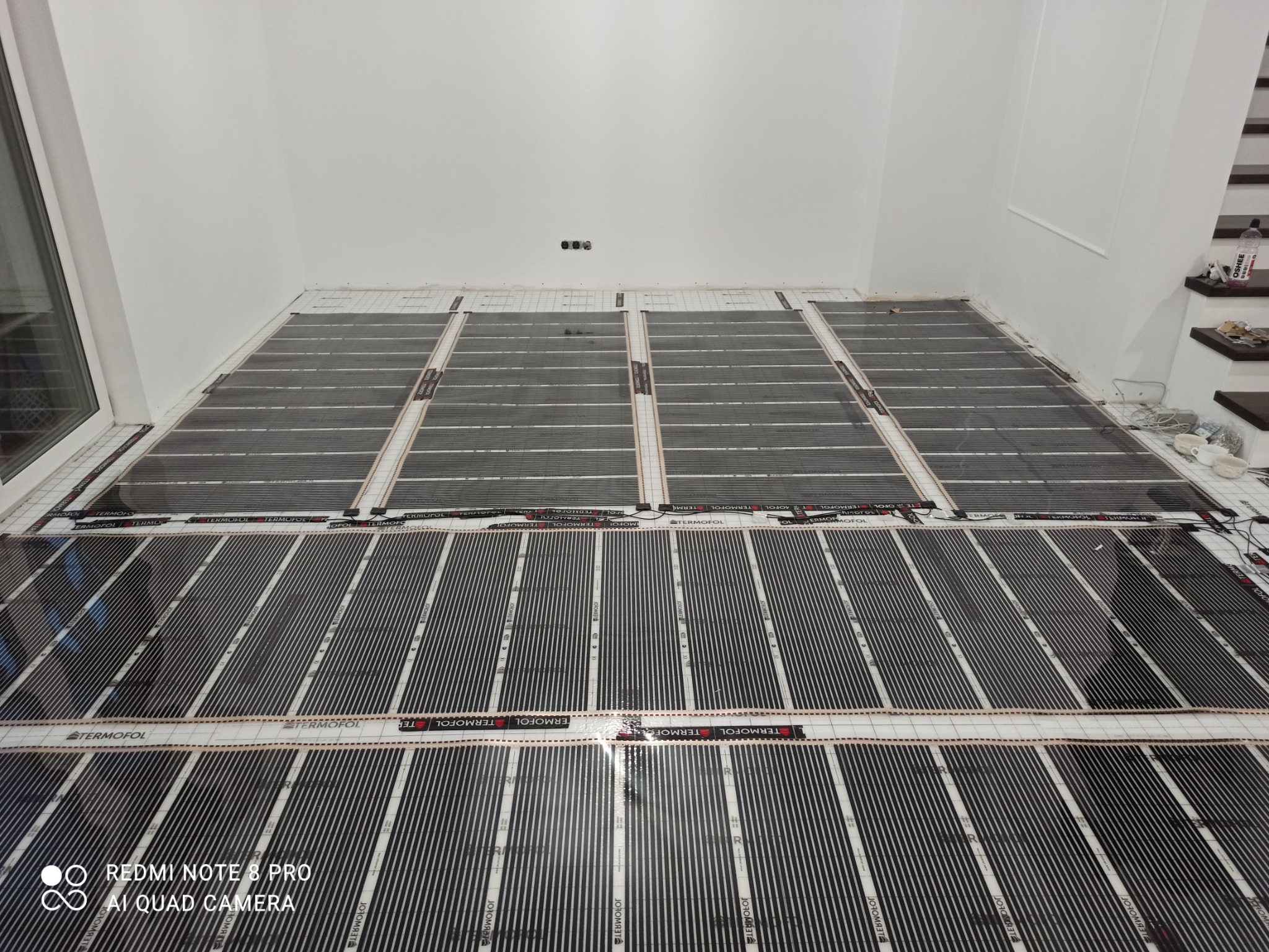 Rozłożone maty grzewcze Termofol na podłodze w jasnym pomieszczeniu, przygotowane do montażu, widoczne przewody i puszki elektryczne w tle.