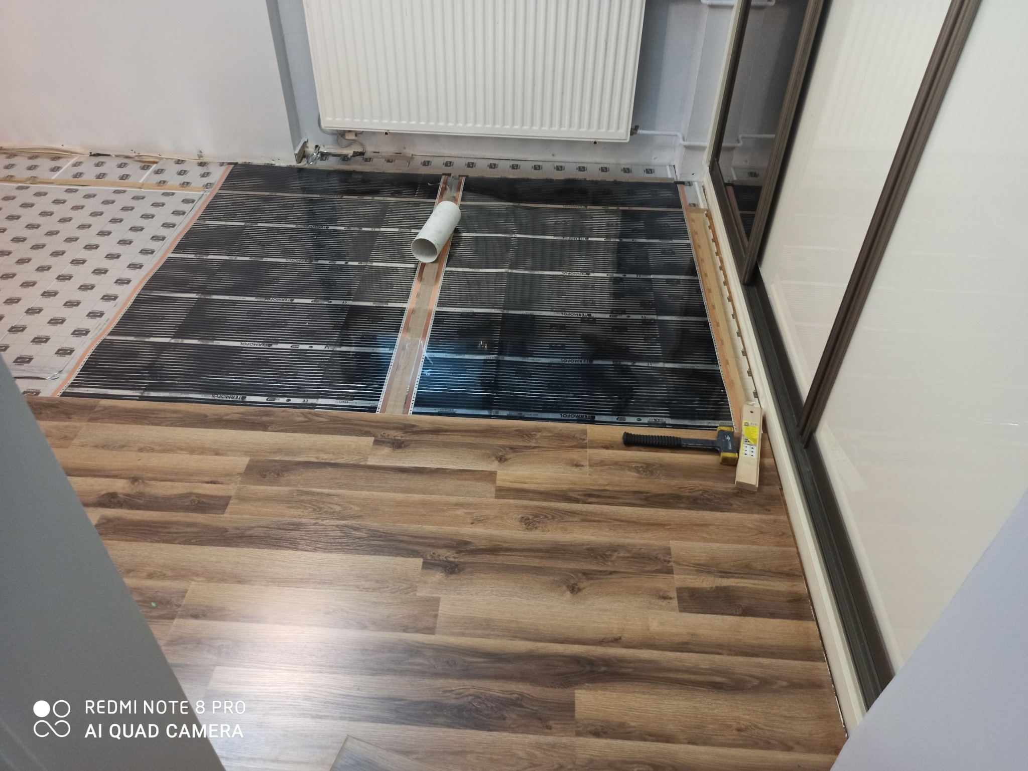Częściowo ułożone maty grzewcze Termofol na podłodze z paneli imitujących drewno, widoczny fragment rury i narzędzia montażowe przy ścianie z grzejnikiem.