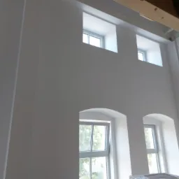 Wnętrze z białymi ścianami, widoczne okna o różnych kształtach (prostokątne i łukowe) oraz elementy drewnianej konstrukcji stropu.