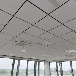 Kasetonowy sufit podwieszany z czarnymi profilami, wbudowaną klimatyzacją i oświetleniem, widok z wnętrza budynku z dużymi oknami na zewnątrz.
