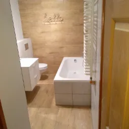 Biała, prostokątna wanna obudowana płytkami w łazience z beżowymi płytkami imitującymi drewno na ścianach i podłodze, widoczna umywalka z szafką i toaleta w tle, dekoracja ścienna 'hello'...