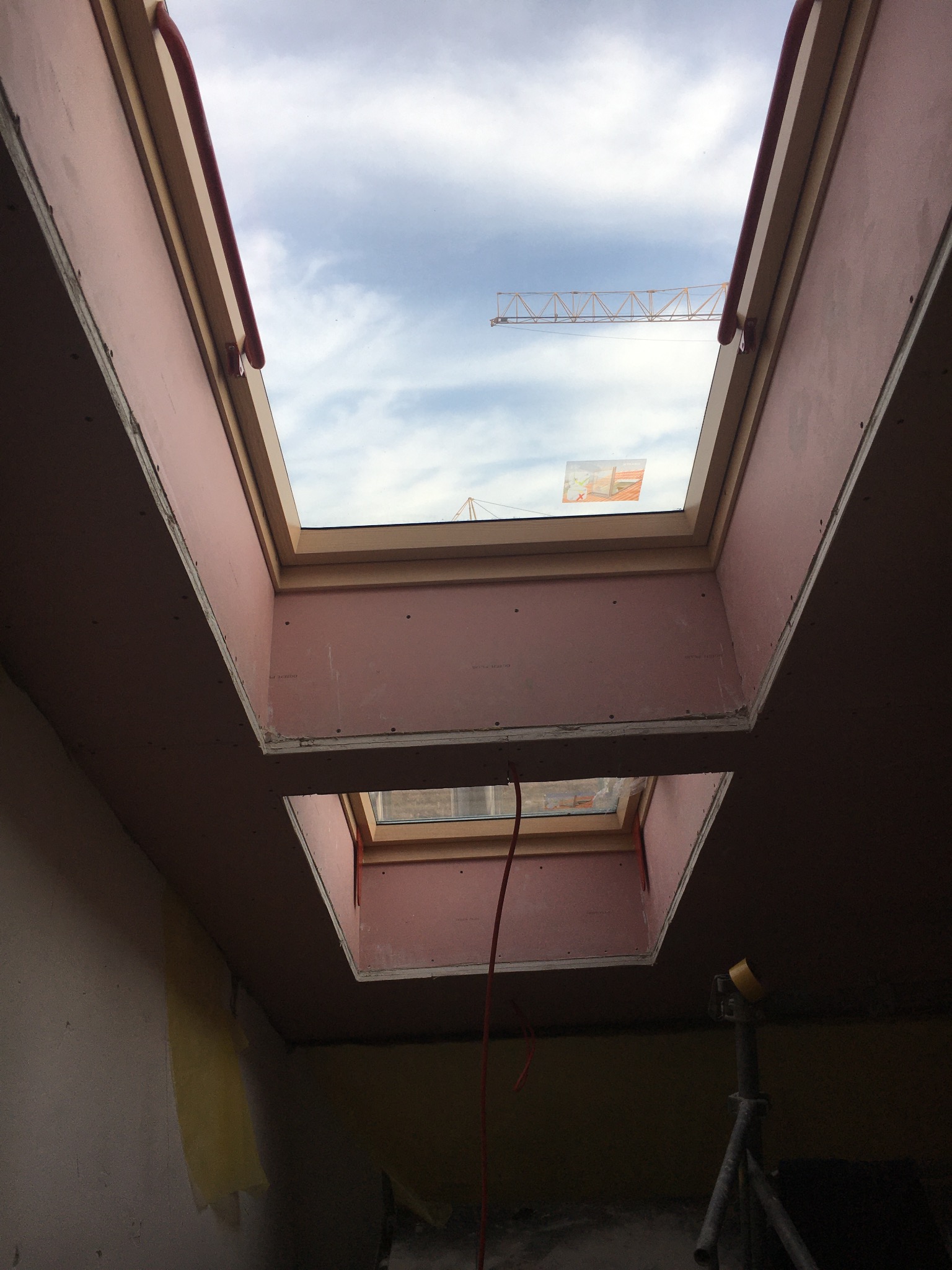 Montaż dwóch okien dachowych w suficie z płyt kartonowo-gipsowych, widoczny dźwig budowlany przez okno.