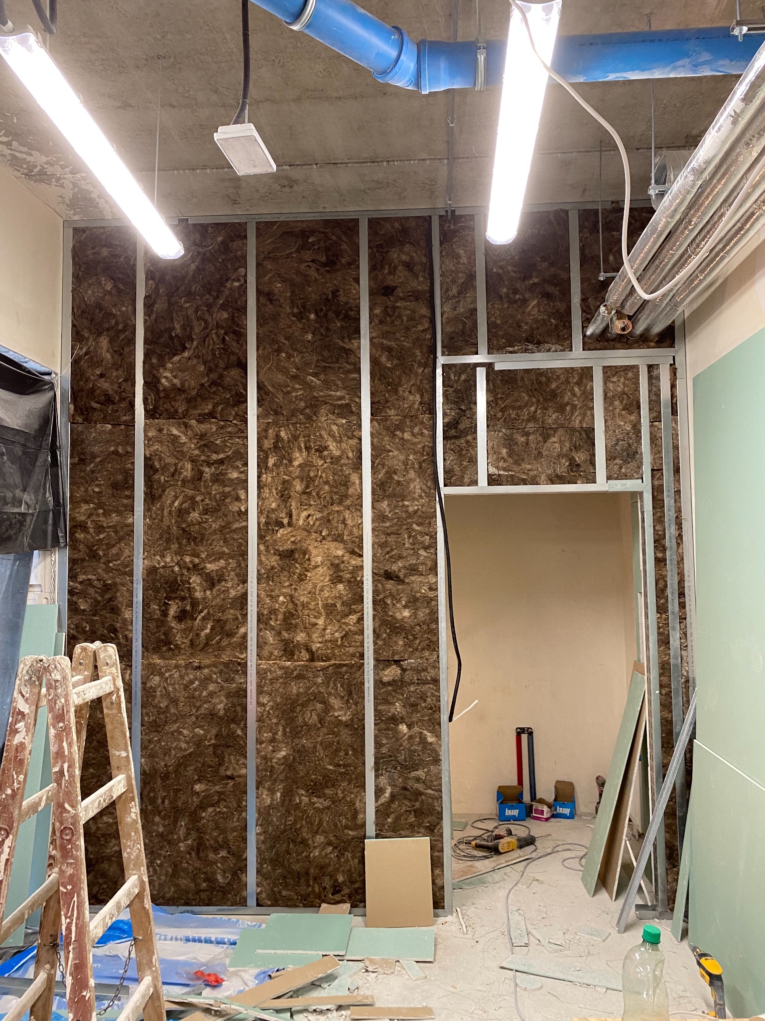 Montaż ściany działowej z wełną mineralną w metalowej konstrukcji, widoczne płyty kartonowo-gipsowe, drabina malarska, narzędzia i materiały budowlane w surowym wnętrzu.