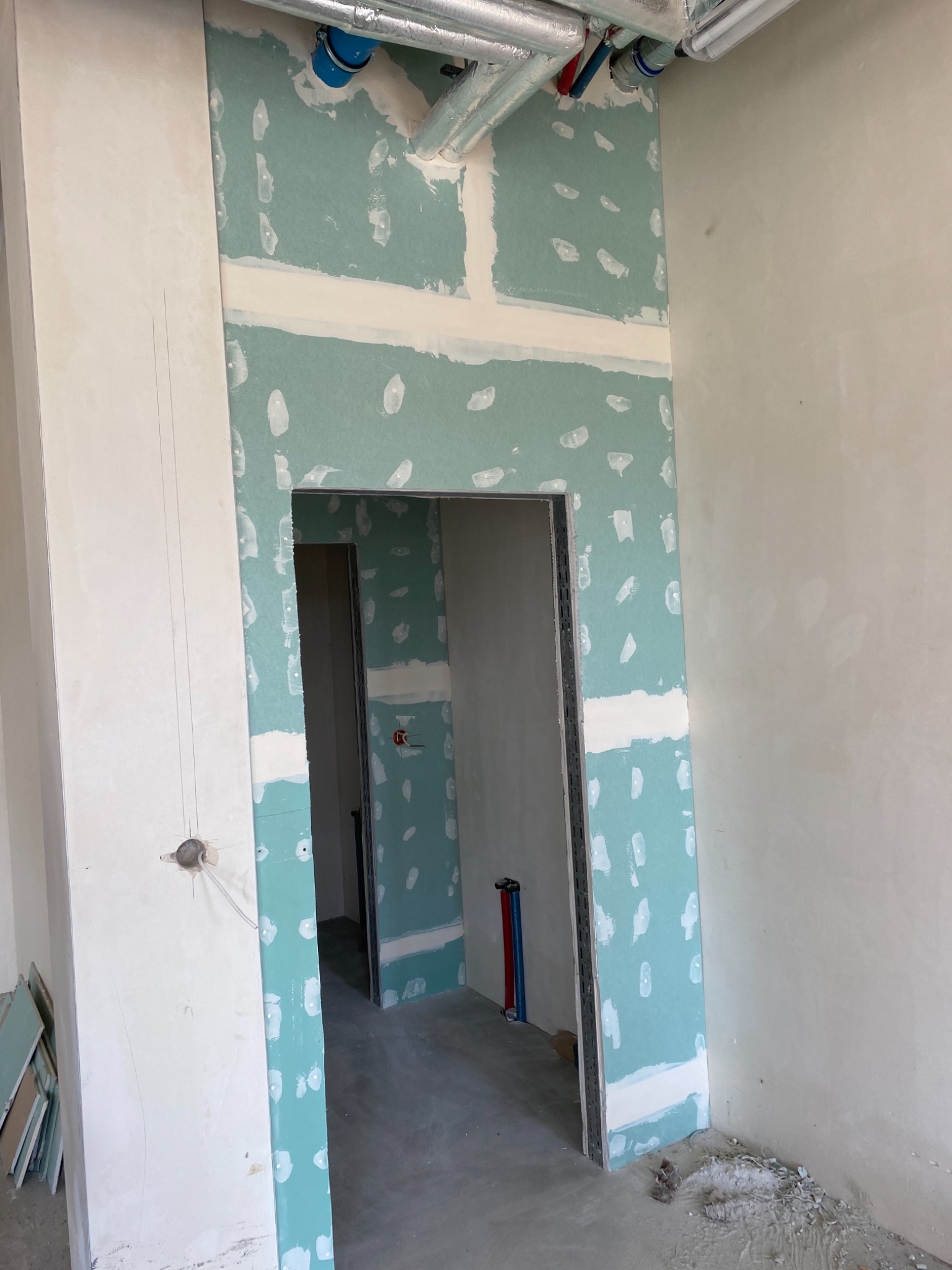 Montaż płyty kartonowo-gipsowej wokół ościeżnicy drzwiowej, widoczne rury instalacji pod sufitem i wystające przewody elektryczne, ślady po szpachlowaniu.